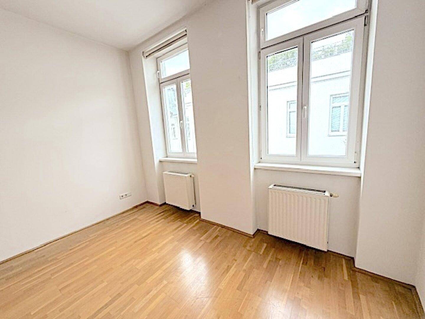 Helles, leeres Schlafzimmer mit Parkettboden und zwei Fenstern.