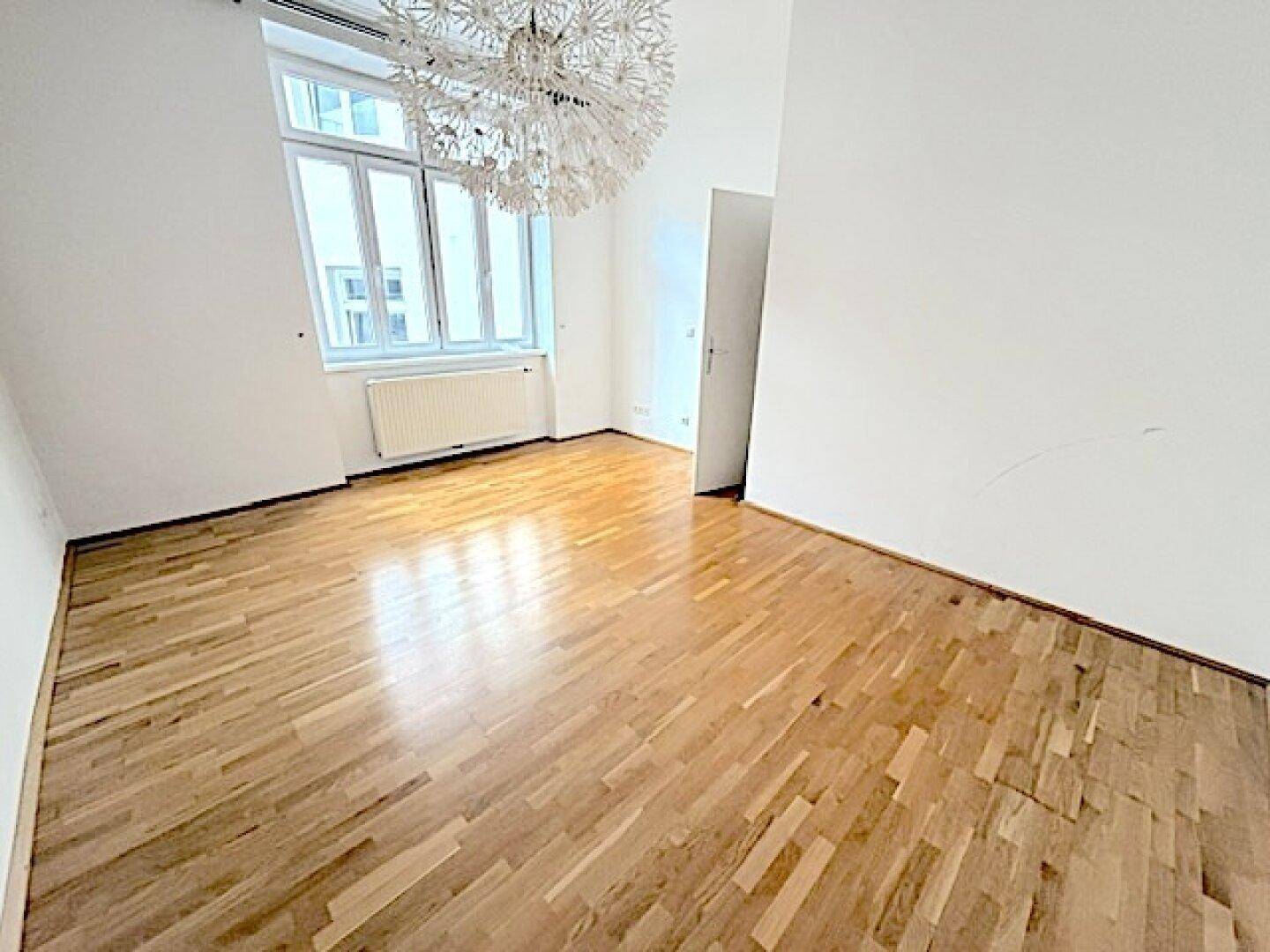 Helles Wohnzimmer mit Parkettboden, großen Fenstern und einer modernen Deckenleuchte.