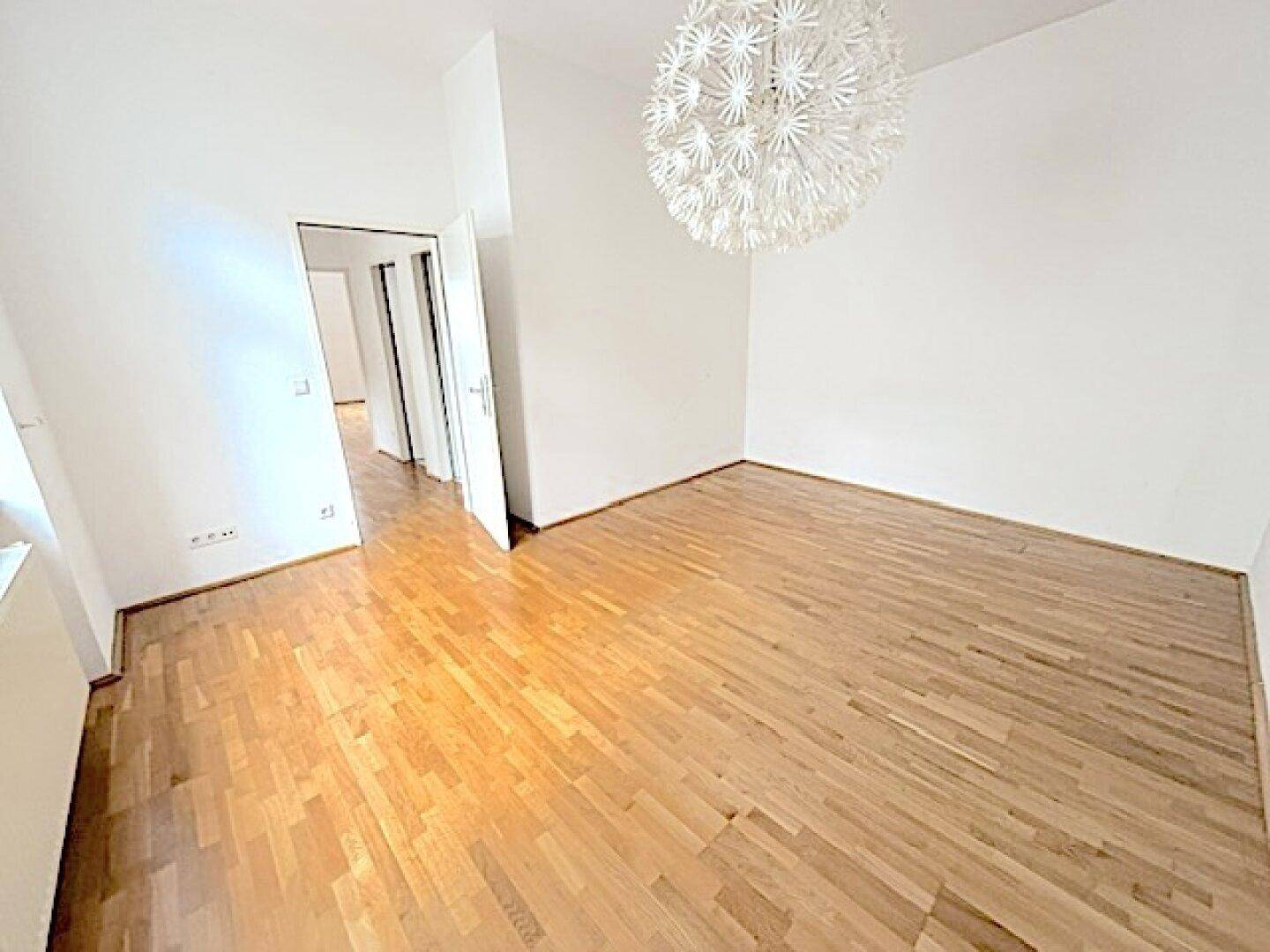 Leerer Raum mit Parkettboden, weißer Deckenleuchte und Zugang zu einem weiteren Bereich.