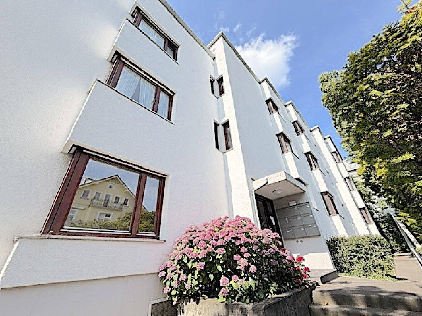 Moderne Wohnhausfassade mit gepflegten Blumenbeeten und Eingangsbereich unter blauem Himmel.