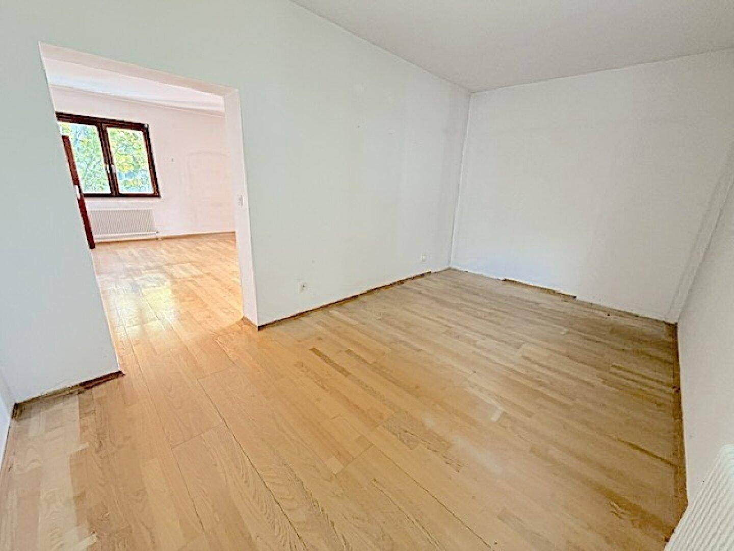 Heller Raum mit Parkettboden, der zu einem weiteren Zimmer mit Fenster führt.