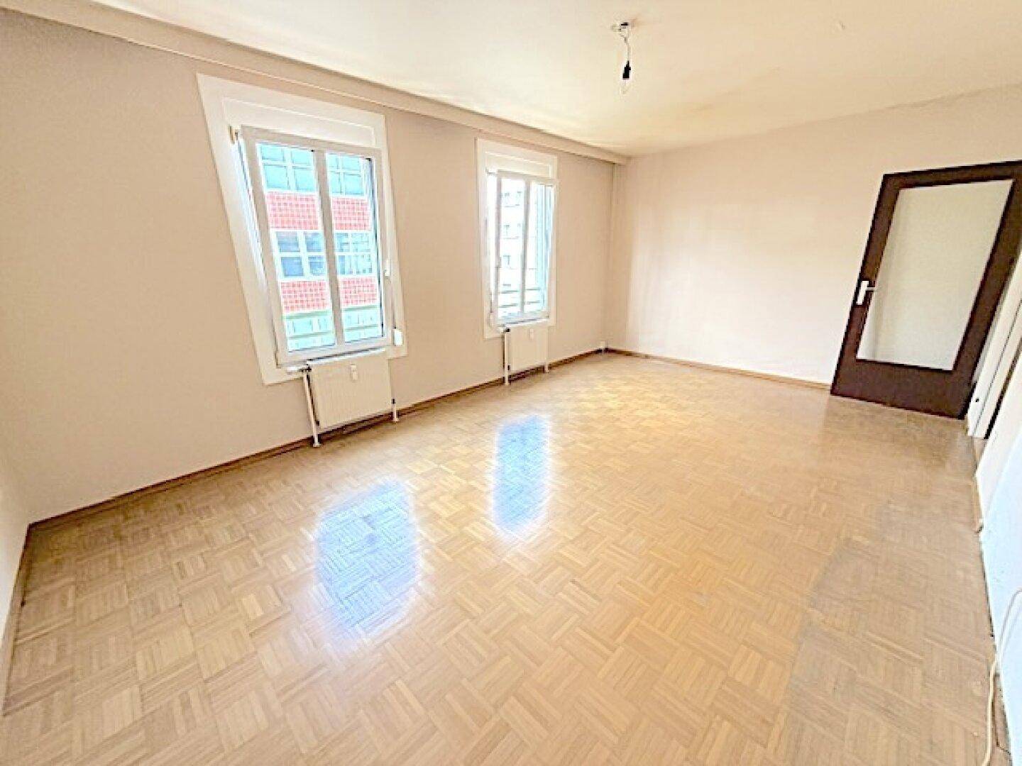 Leeres Zimmer mit Fischgrätparkett und zwei Fenstern, bereit zur individuellen Gestaltung.
