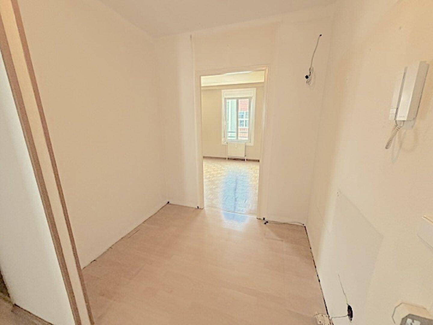 Heller Flur mit hellem Boden, der zu einem weiteren Zimmer mit Fenster führt.