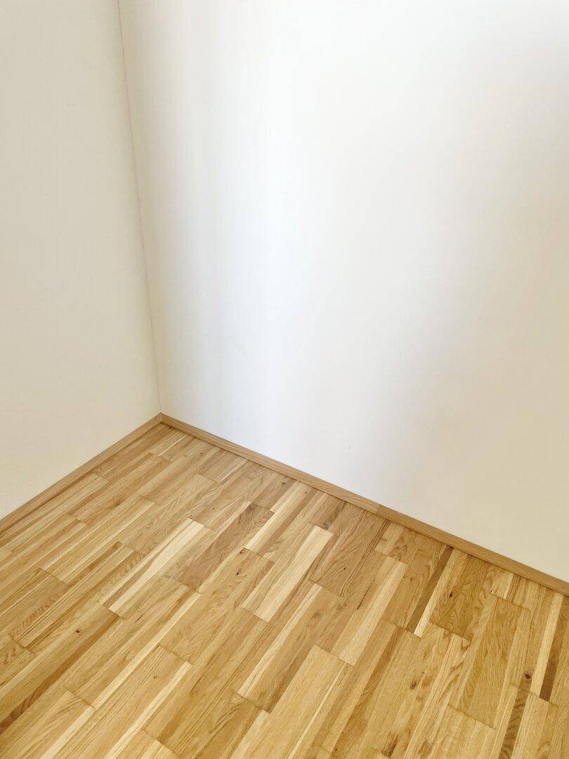 Detailansicht einer Raumecke mit hellem Parkettboden und sauberen weißen Wänden.