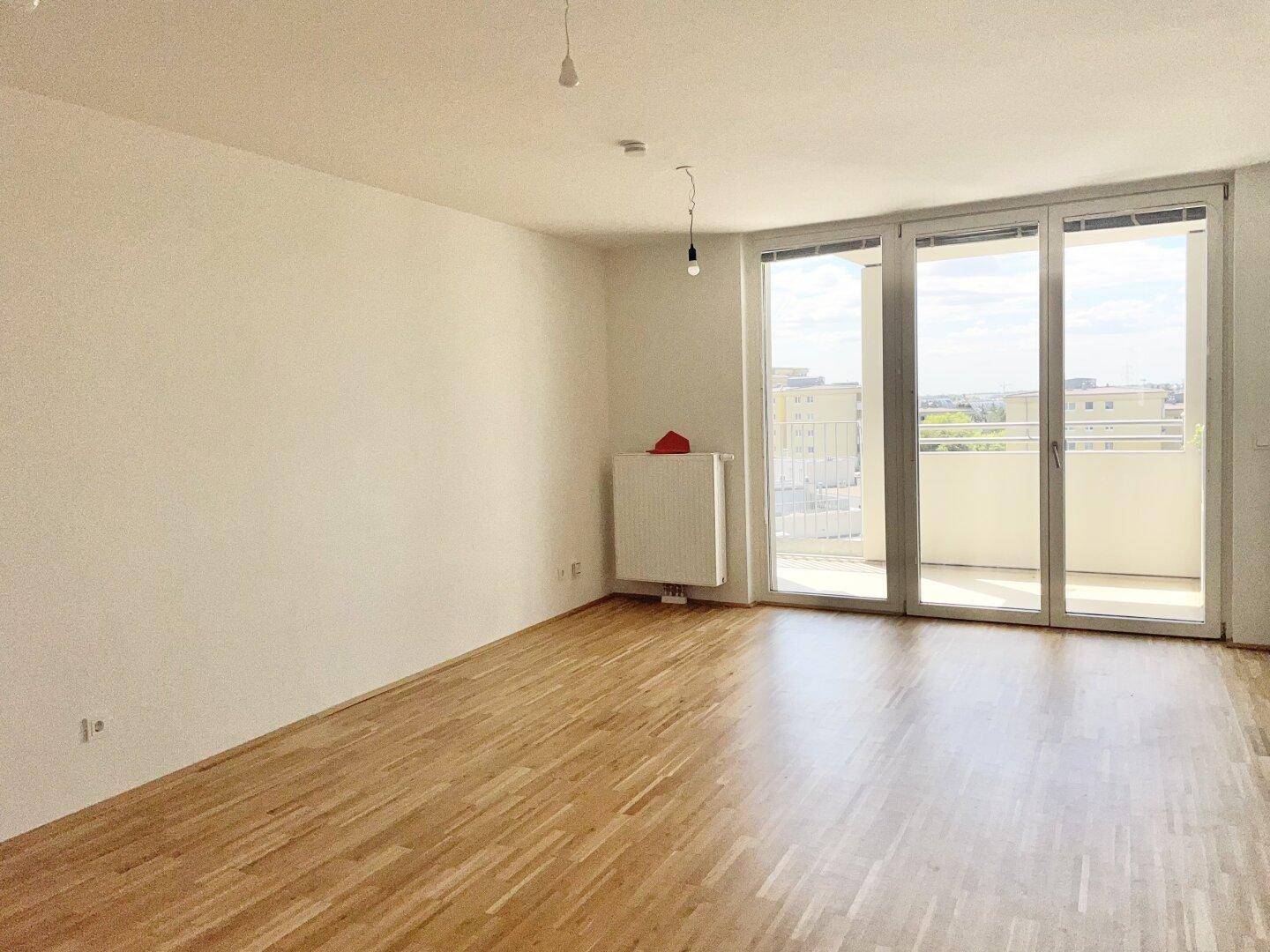 Heller, leerer Wohnbereich mit Parkettboden und großen Fenstern, die Zugang zu einem Balkon bieten.