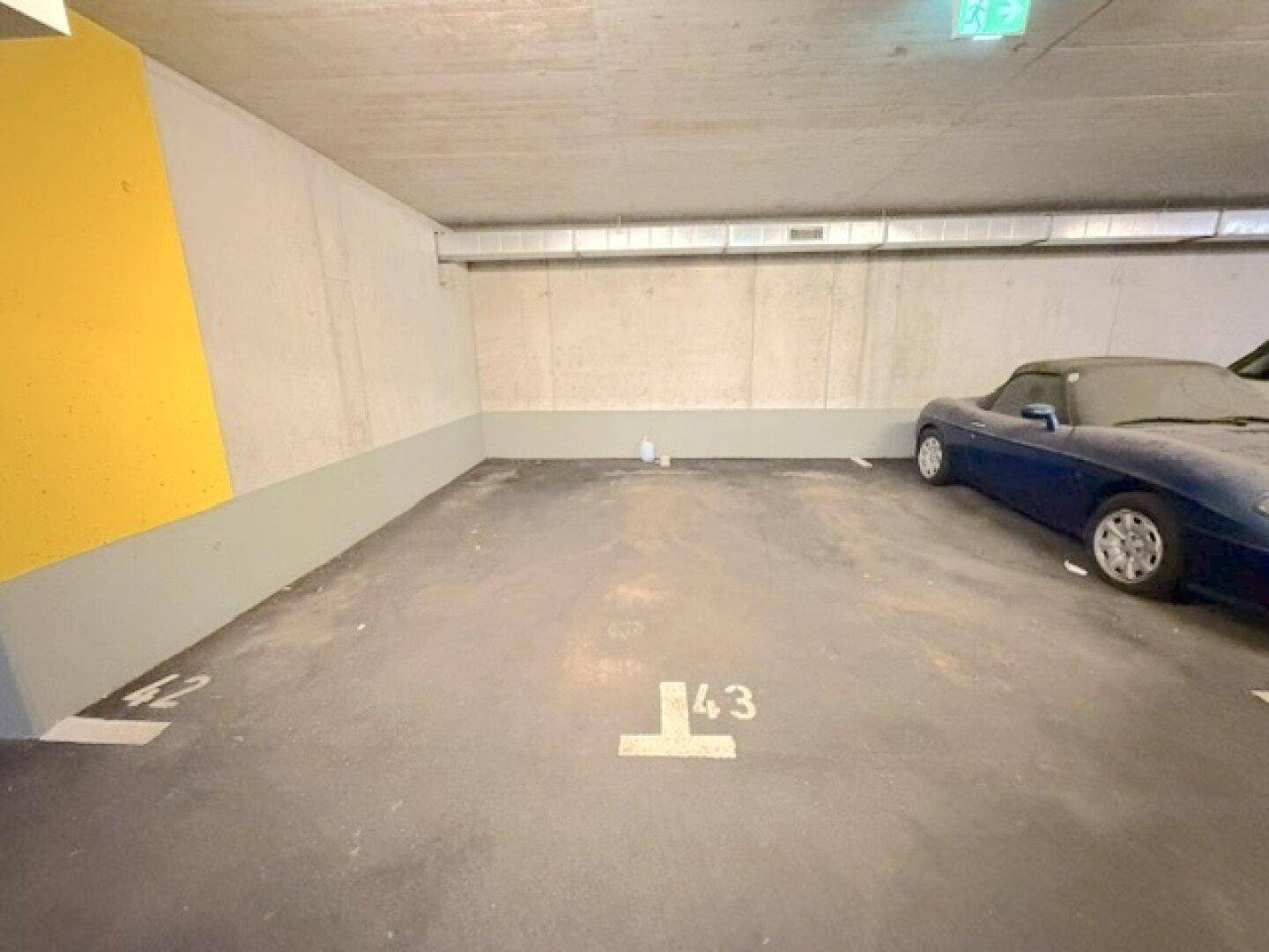 Leere Tiefgaragenstellplätze mit Betonboden und gelbem Pfeiler, ein Sportwagen parkt daneben.