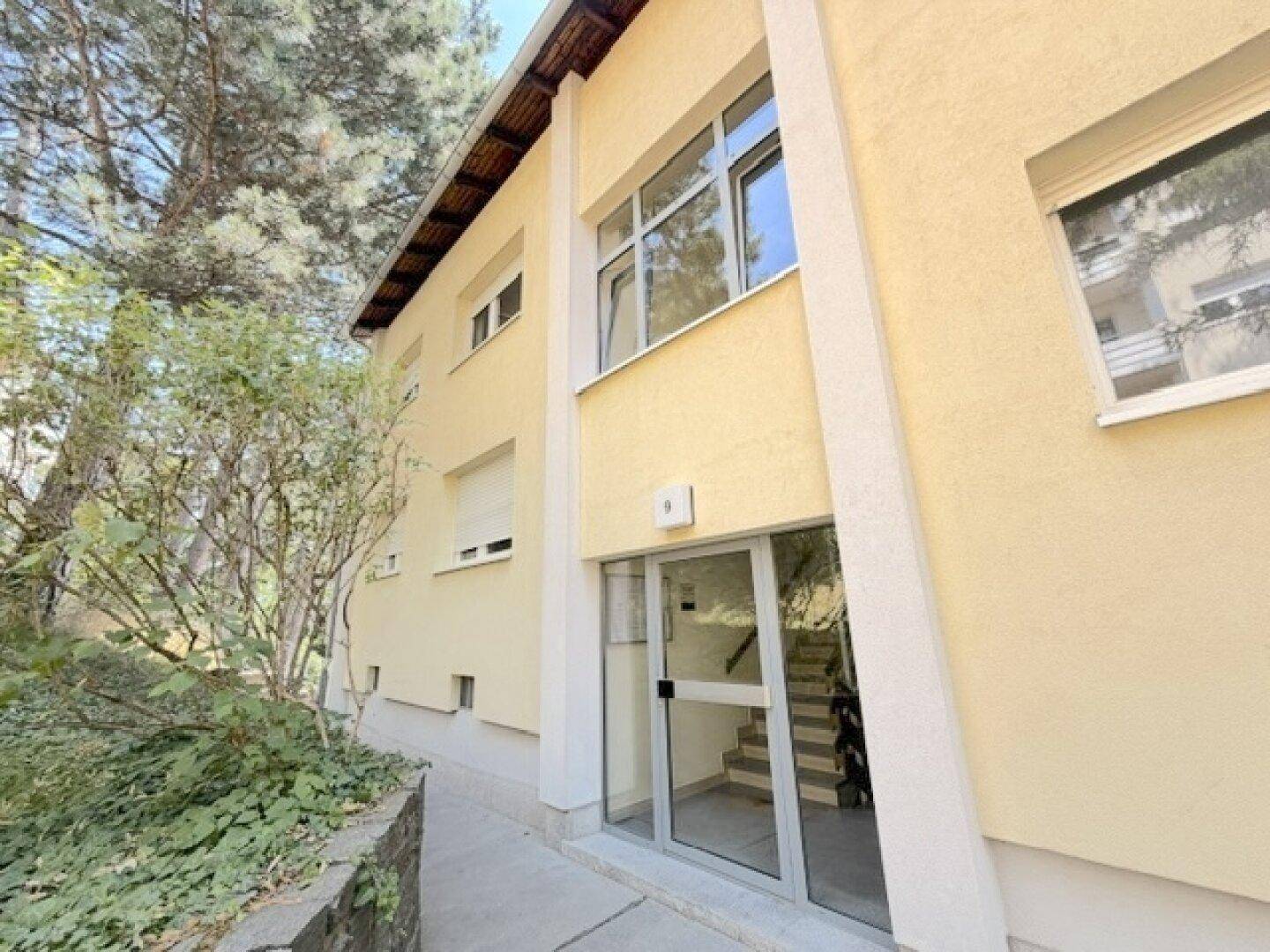 Eingangsbereich eines Mehrfamilienhauses mit Glasfront und Treppenhaus im Inneren.