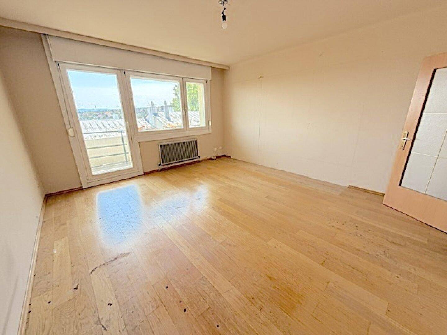 Helles, leeres Zimmer mit Holzboden und Fensterfront mit Blick ins Grüne.