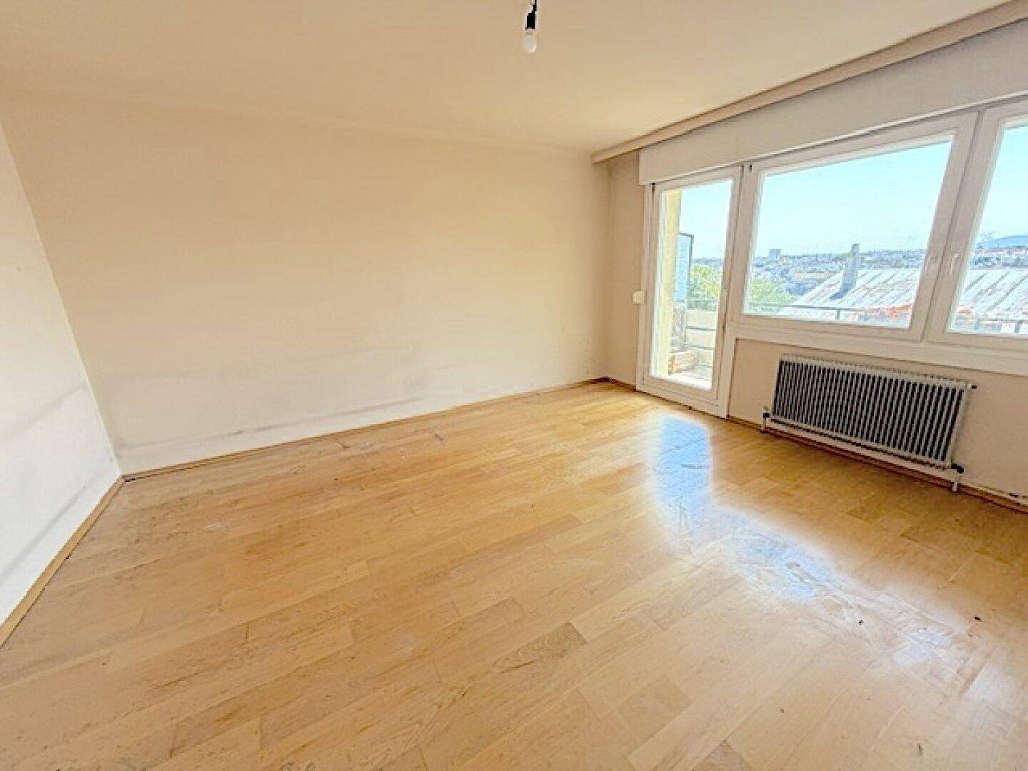 Leeres Zimmer mit Holzboden, großen Fenstern und Zugang zu einem Balkon.