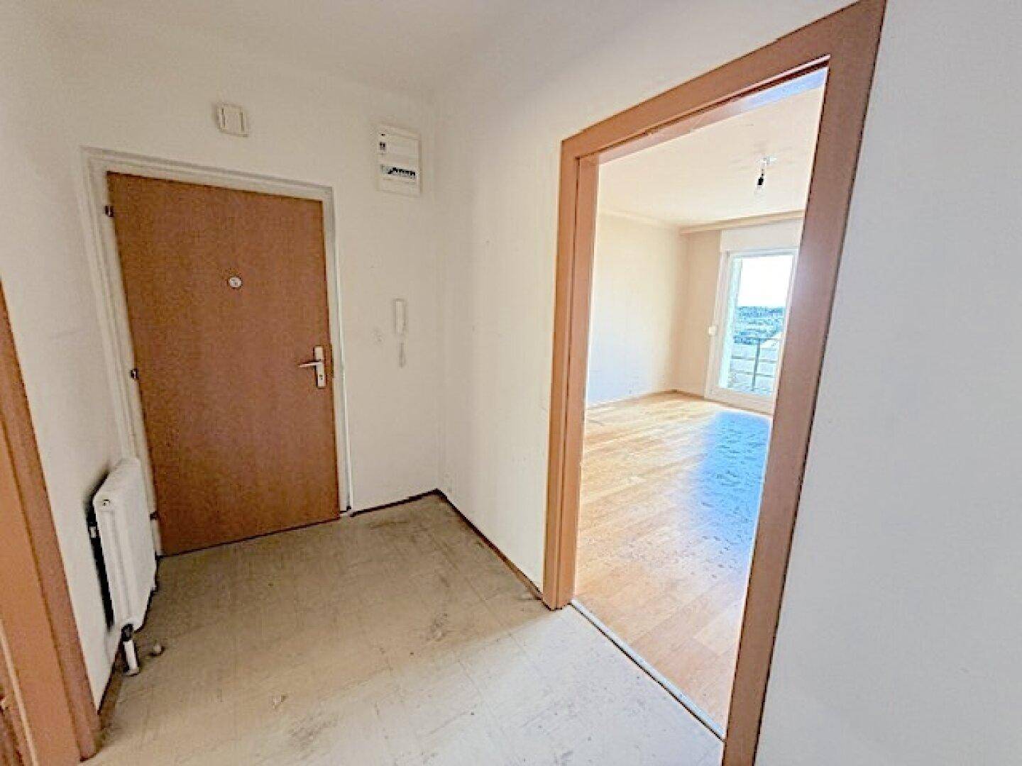 Flur mit Eingangstür und Zugang zu einem hellen Zimmer mit Holzboden.