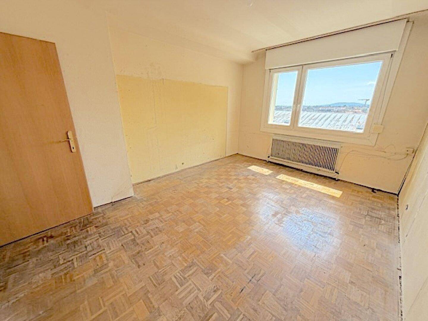 Leeres Zimmer mit Parkettboden, Fenster und viel Tageslicht.