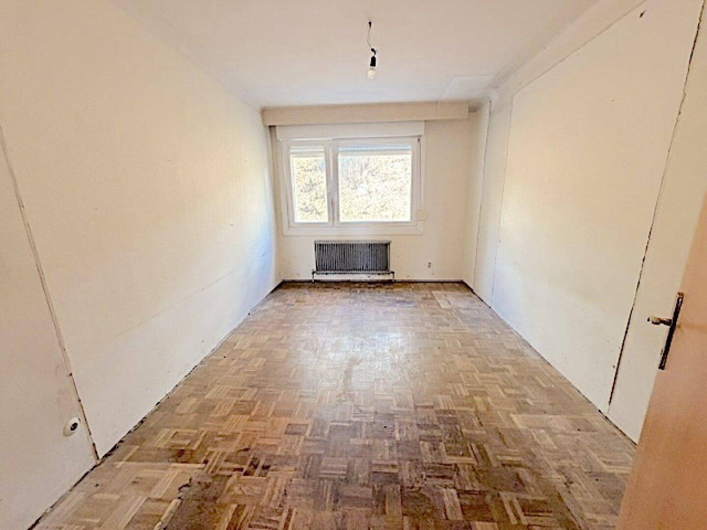 Leeres Schlafzimmer mit Fischgrätparkett, Fenster und Heizkörper.
