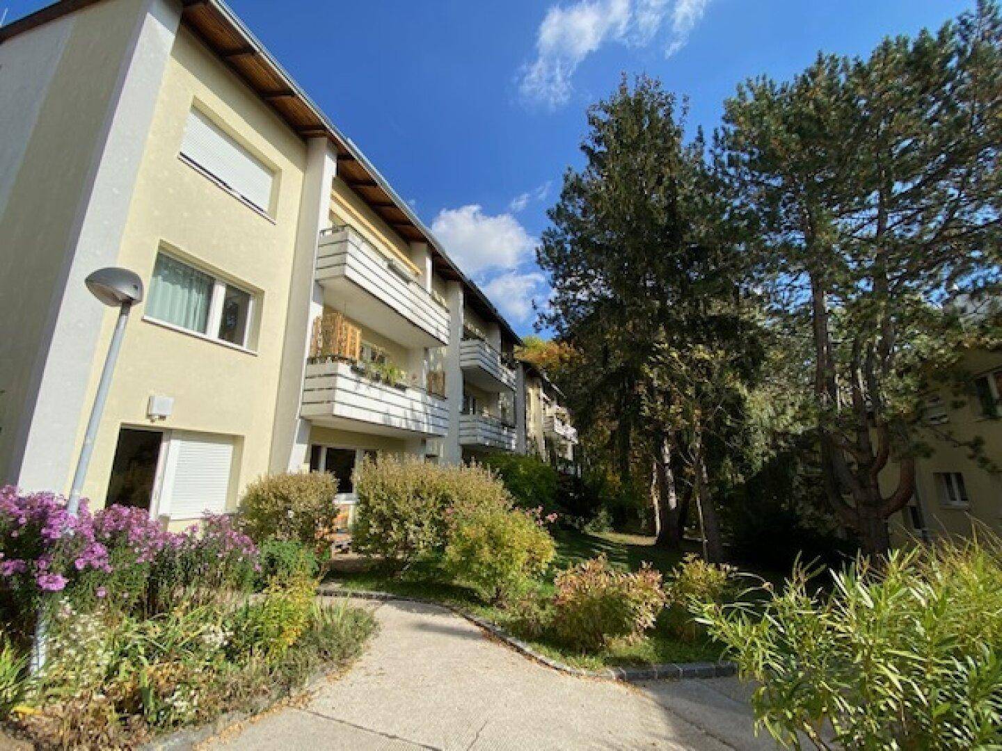 Mehrfamilienhaus mit mehreren Balkonen und Rollläden, umgeben von Bäumen und Sträuchern unter blauem Himmel.