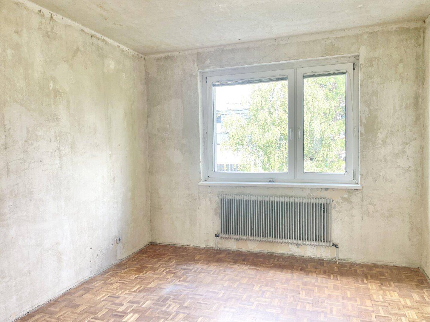 Leeres Zimmer mit Parkettboden und Fenster, das viel Tageslicht hereinlässt.