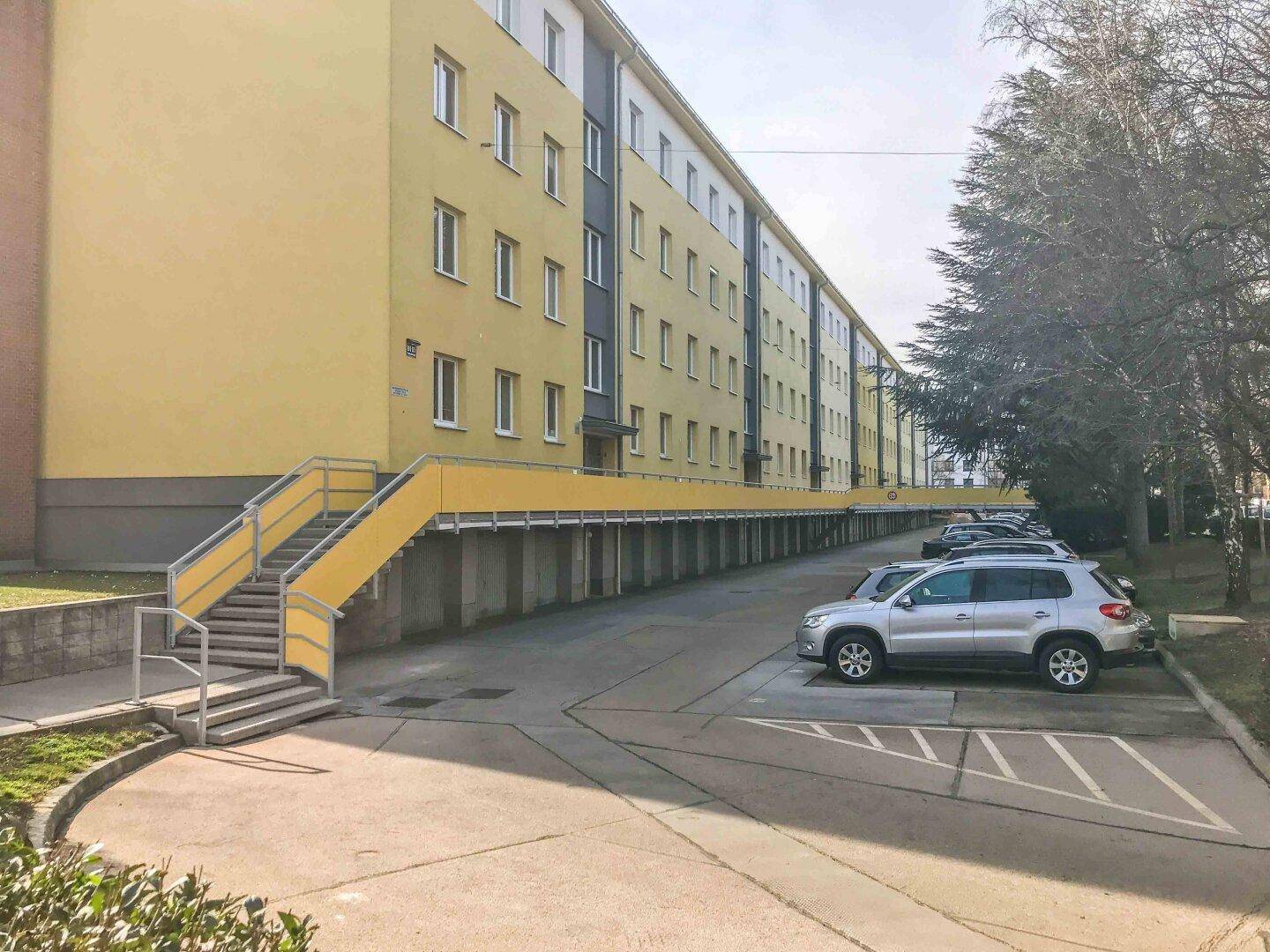 Außenansicht eines Mehrfamilienhauses mit gelber Fassade, Treppen und Parkplätzen.
