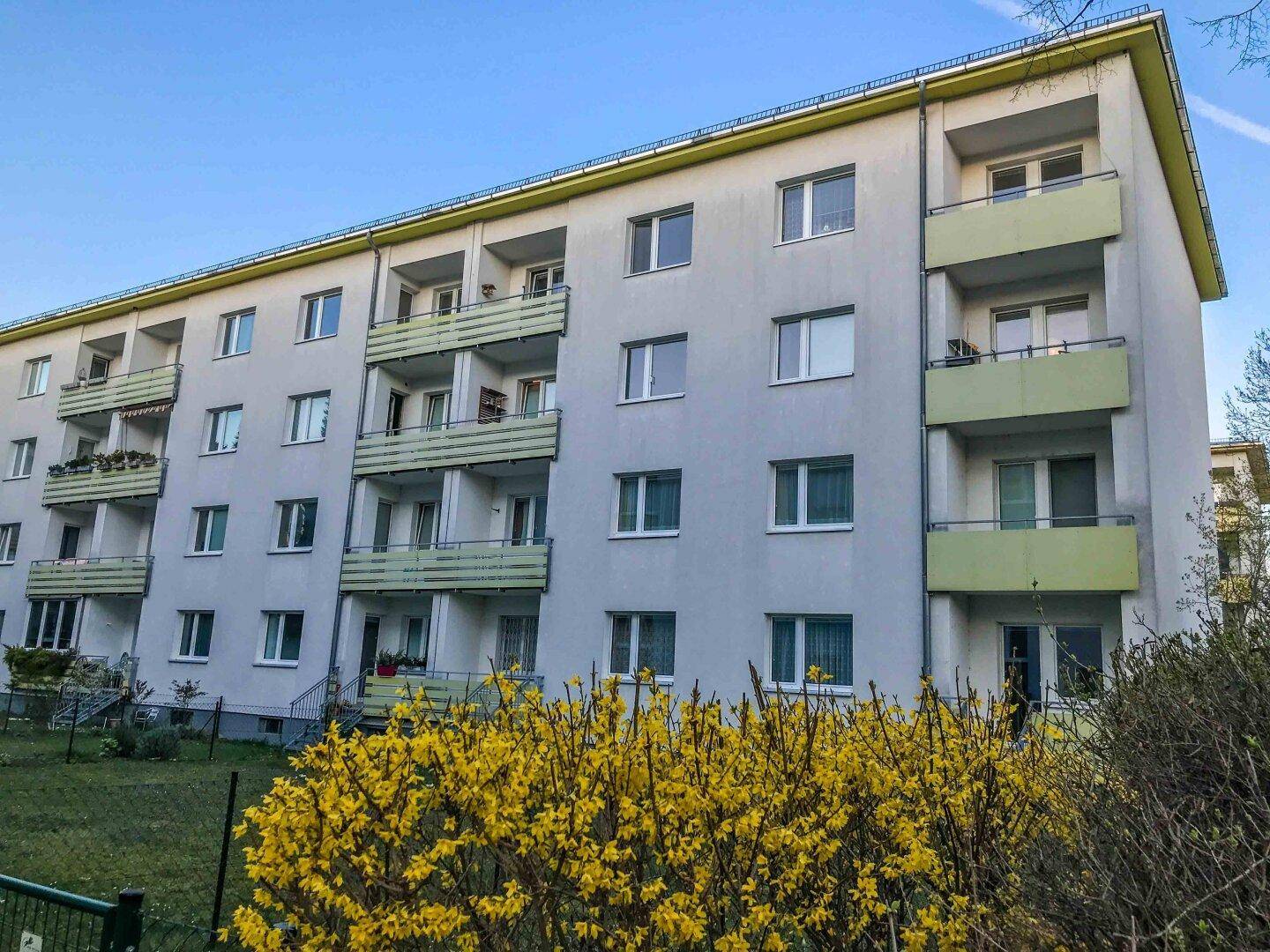 Mehrfamilienhaus mit grauer Fassade und grünen Balkonen, umgeben von blühenden Sträuchern.