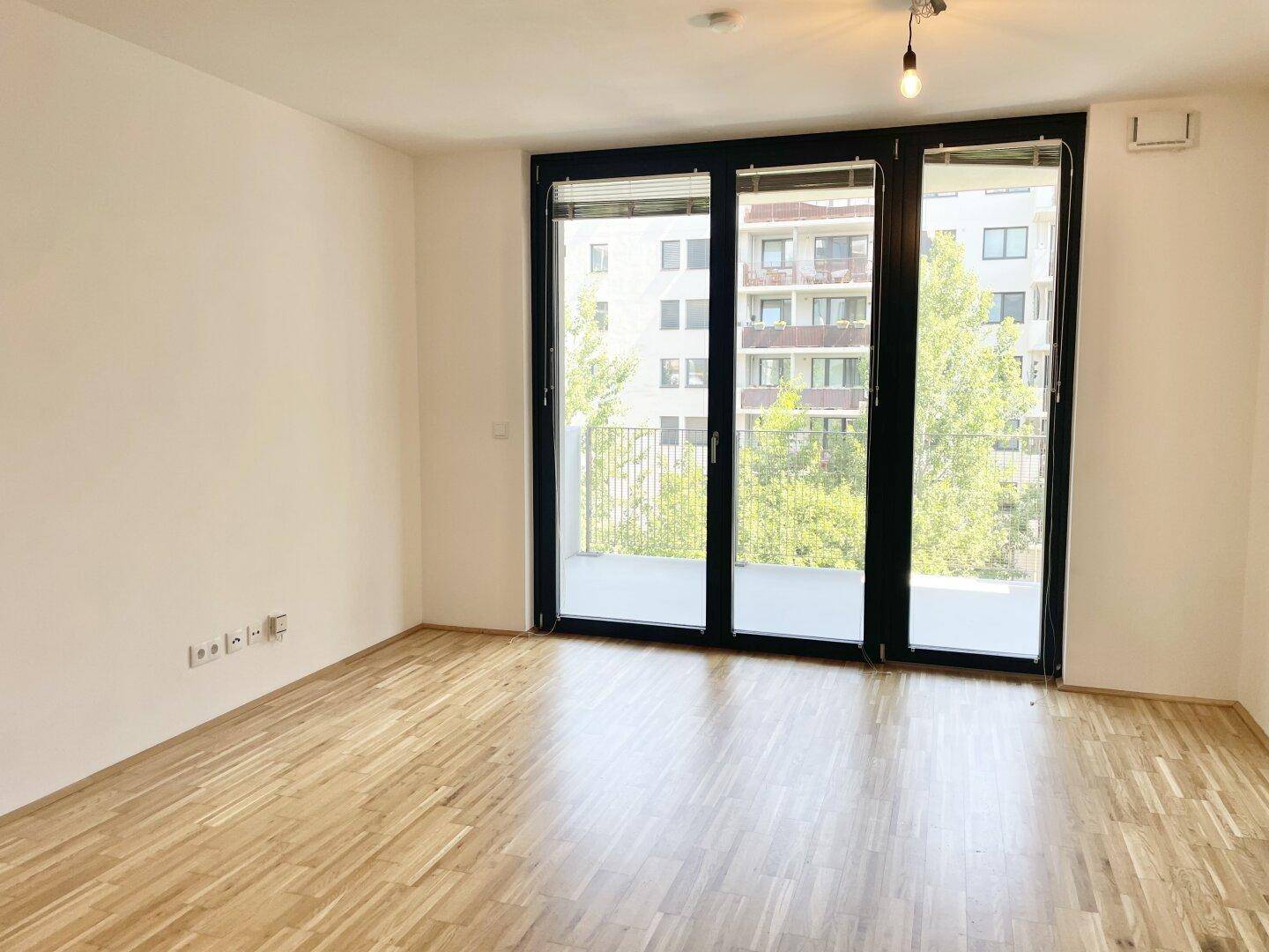 Leeres Wohnzimmer mit Holzboden und großen Fenstern, die viel Tageslicht hereinlassen.