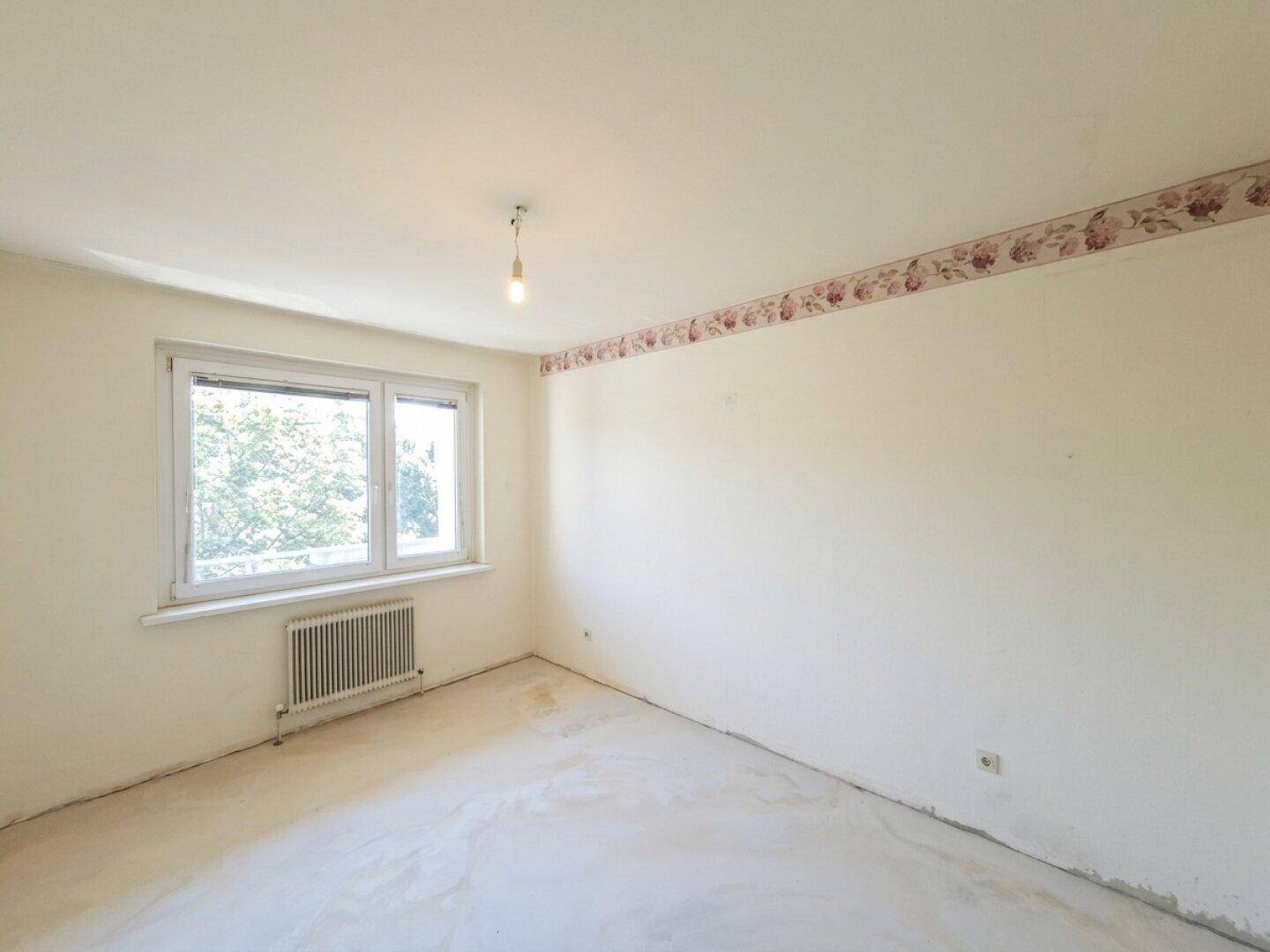 Unmöbliertes Zimmer mit heller Tapete, floraler Bordüre und Fenster mit Heizkörper.