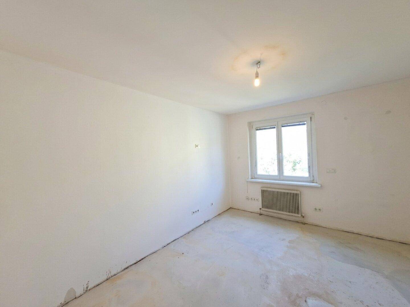 Helles, unmöbliertes Zimmer mit weißen Wänden, Betonboden und Fenster.