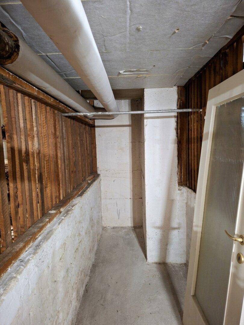 Kellergang mit Betonboden, sichtbaren Rohren und Holzlattenwänden.