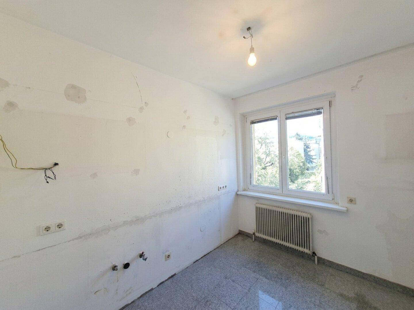 Unmöblierte Küche mit weißen Wänden, grauem Fliesenboden und Fenster.