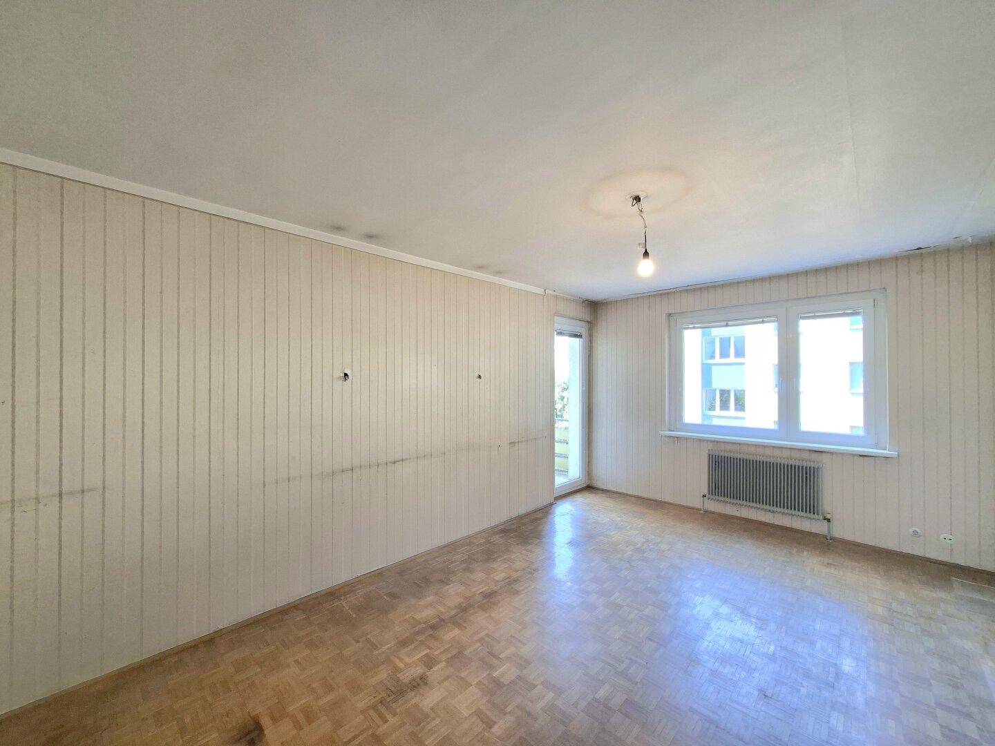 Leeres Wohnzimmer mit Fischgrätparkett, Tapetenresten und Fensterblick ins Grüne.