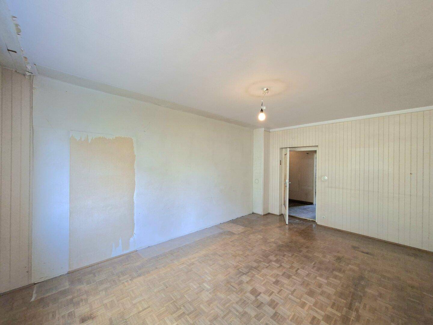 Unmöbliertes Wohnzimmer mit Parkettboden, teilweise entfernter Tapete und Tür zu einem weiteren Raum.