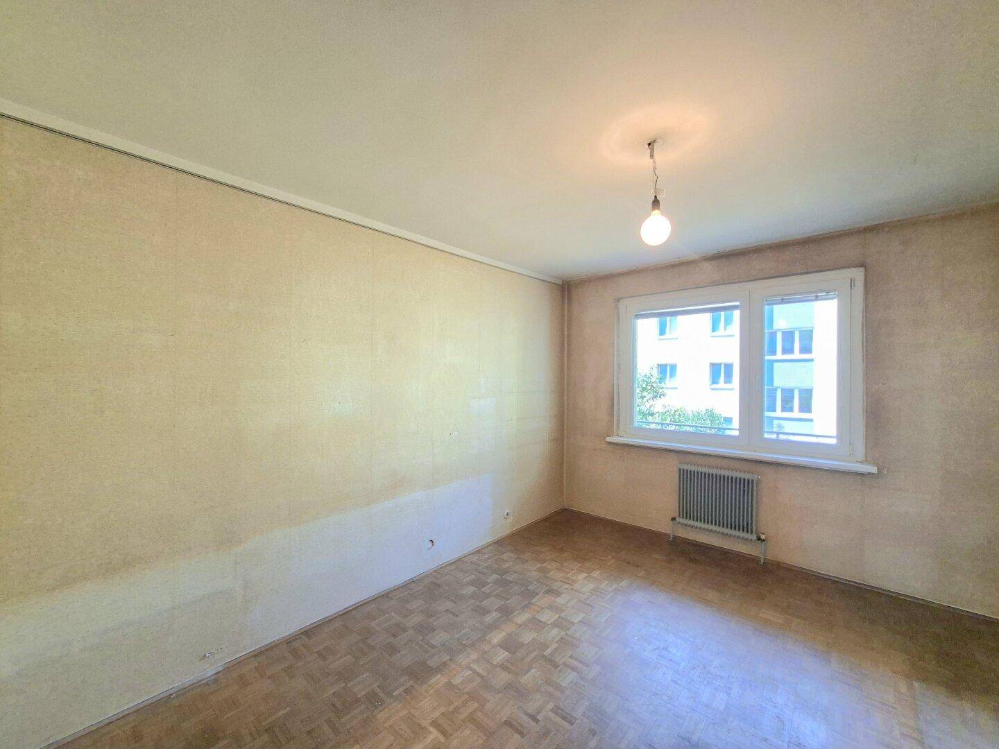 Leeres Zimmer mit Parkettboden, teilweise entfernter Tapete und Fenster mit Blick ins Grüne.