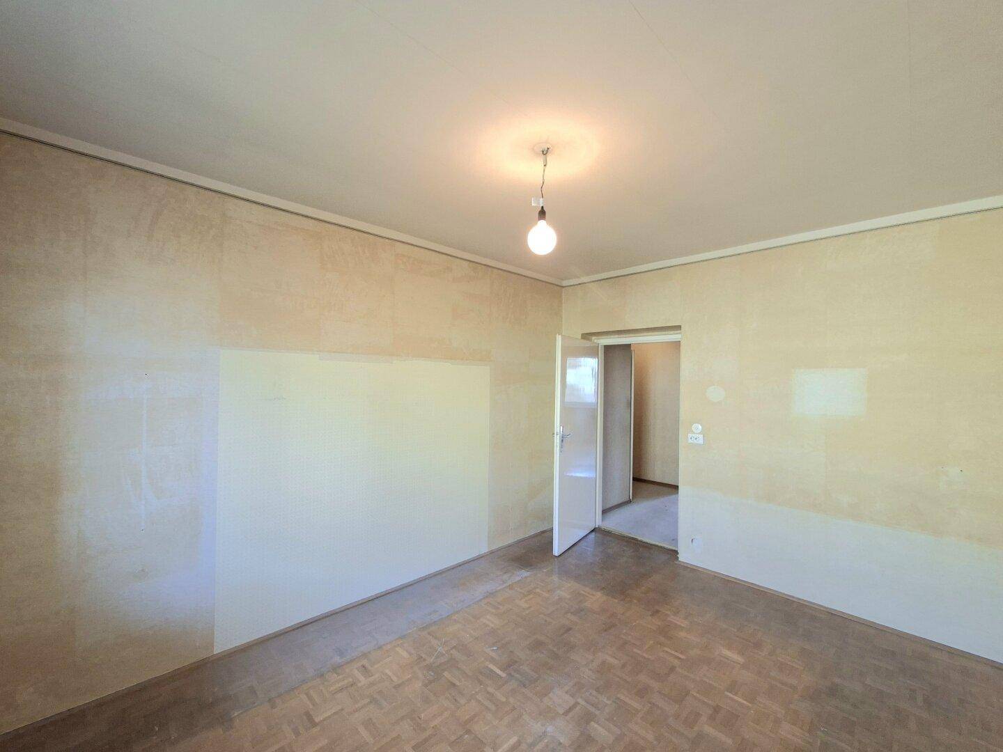 Unmöbliertes Zimmer mit Parkettboden, Tapetenresten und offener Tür zu einem weiteren Raum.