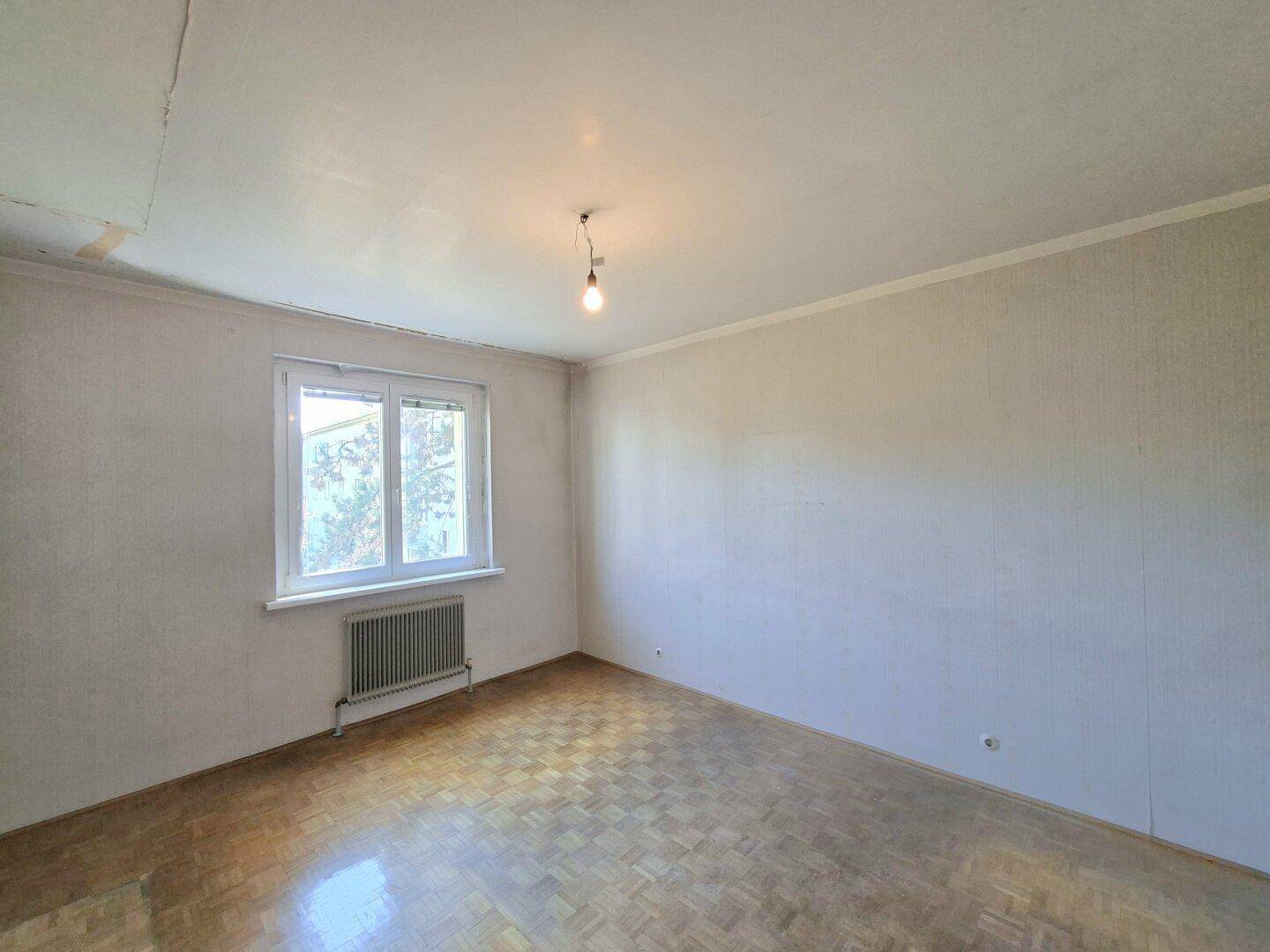 Leeres Zimmer mit Parkettboden, hellen Wänden und Fenster mit Blick auf Bäume.