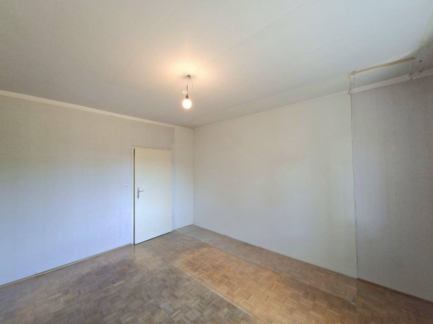 Leeres Zimmer mit Parkettboden, weißen Wänden und einer geschlossenen Tür.