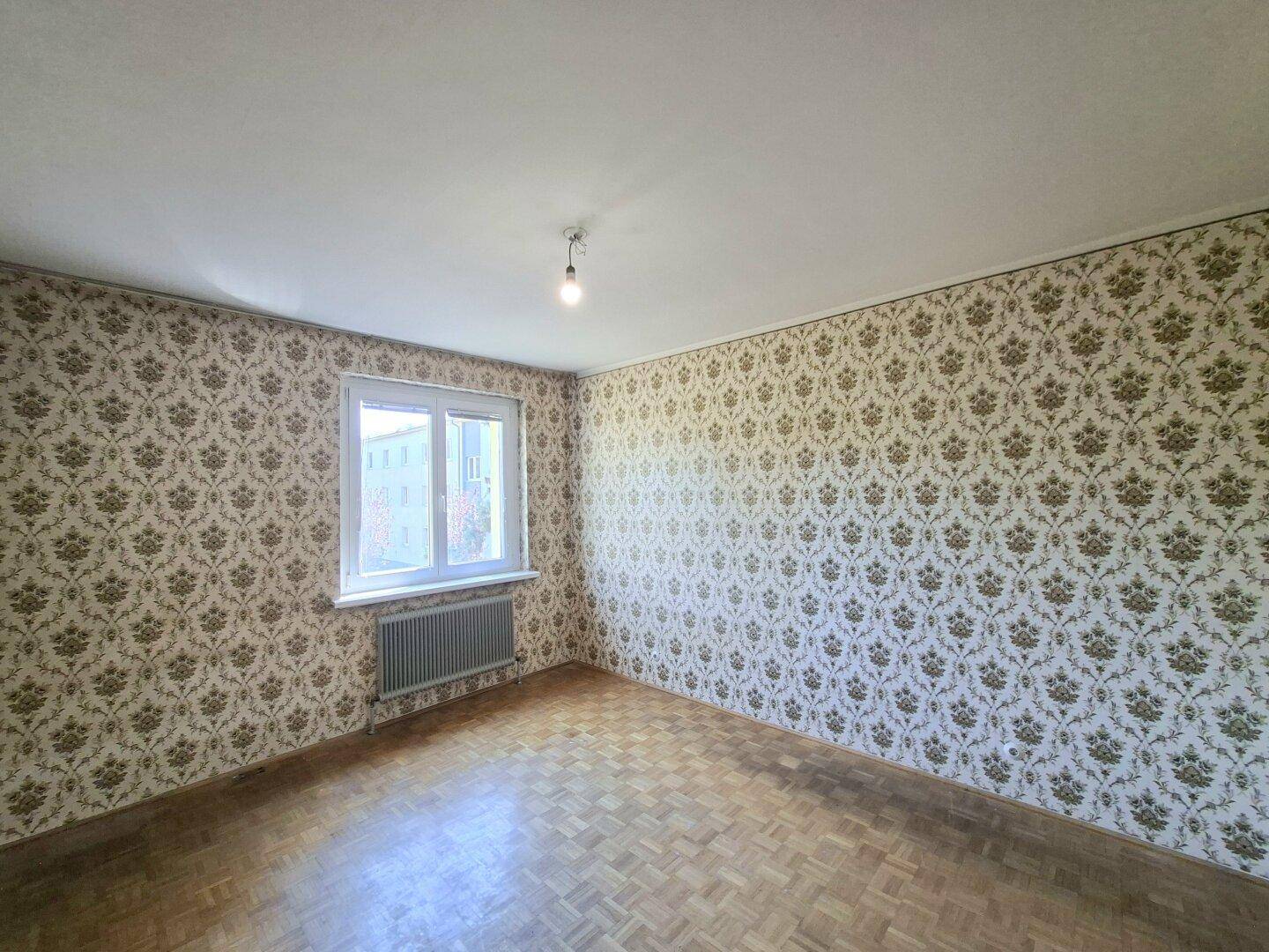 Zimmer mit gemusterten Tapeten, Parkettboden und Fenster mit Blick auf die Umgebung.