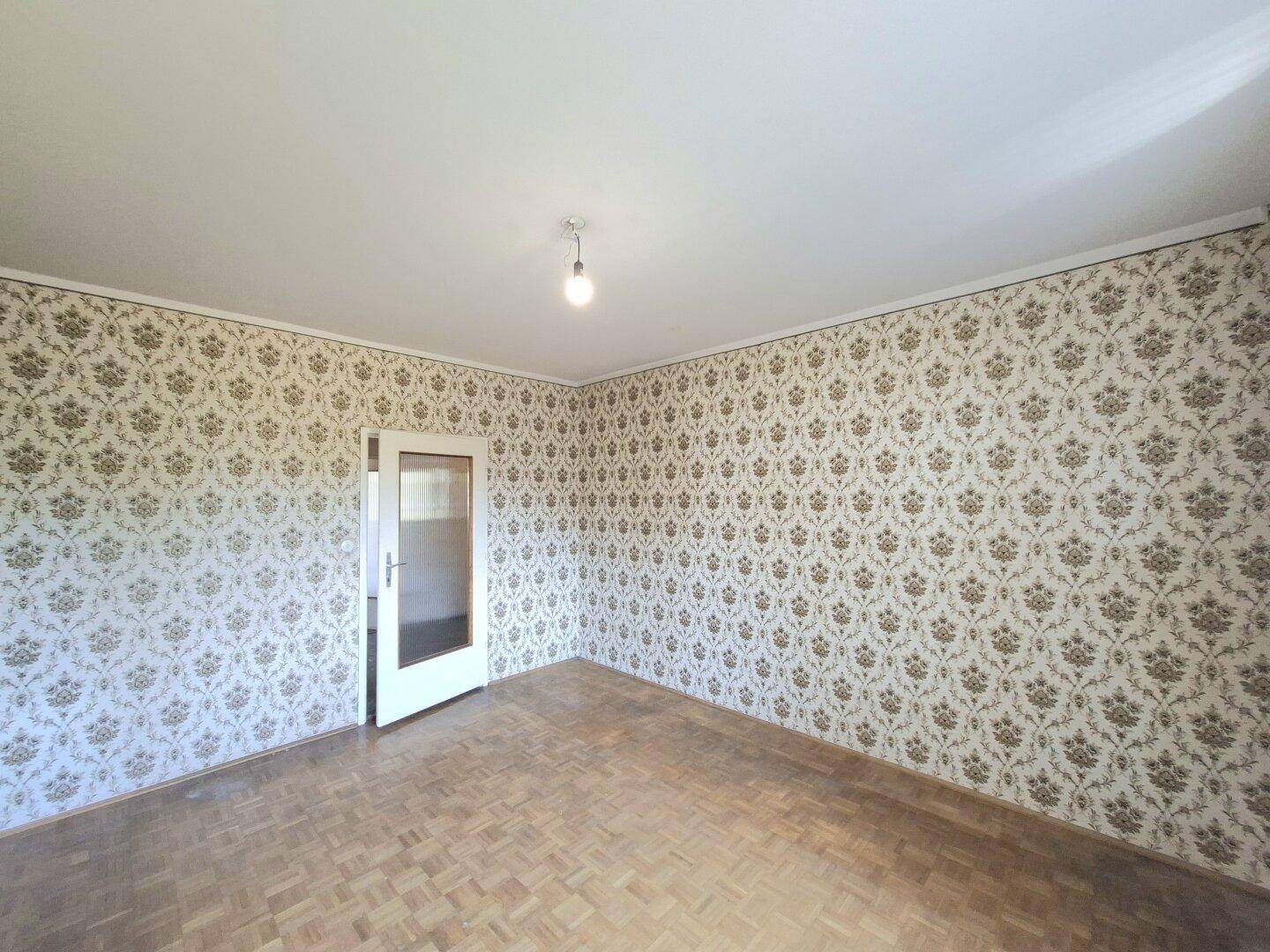 Zimmer mit gemusterten Tapeten, Parkettboden und einer Tür mit Glaseinsatz.