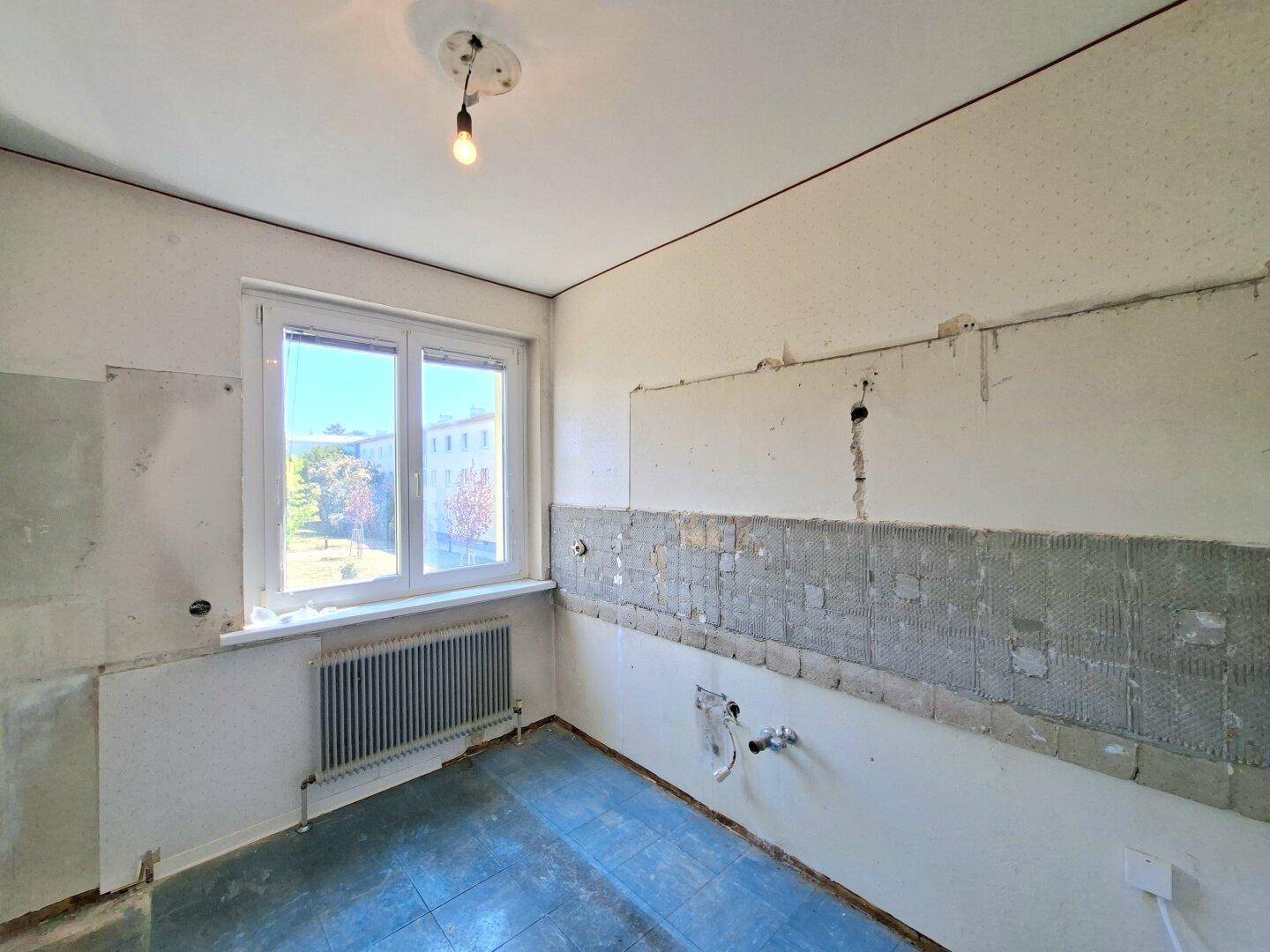 Unmöblierte Küche mit Fliesenboden, entfernten Wandfliesen und Fenster mit Blick ins Grüne.