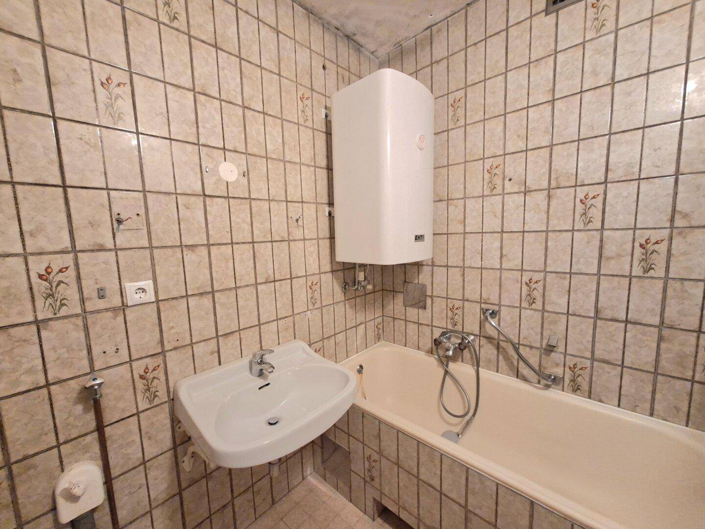 Badezimmer mit beigefarbenen Fliesen, Waschbecken, Badewanne und Warmwasserboiler.