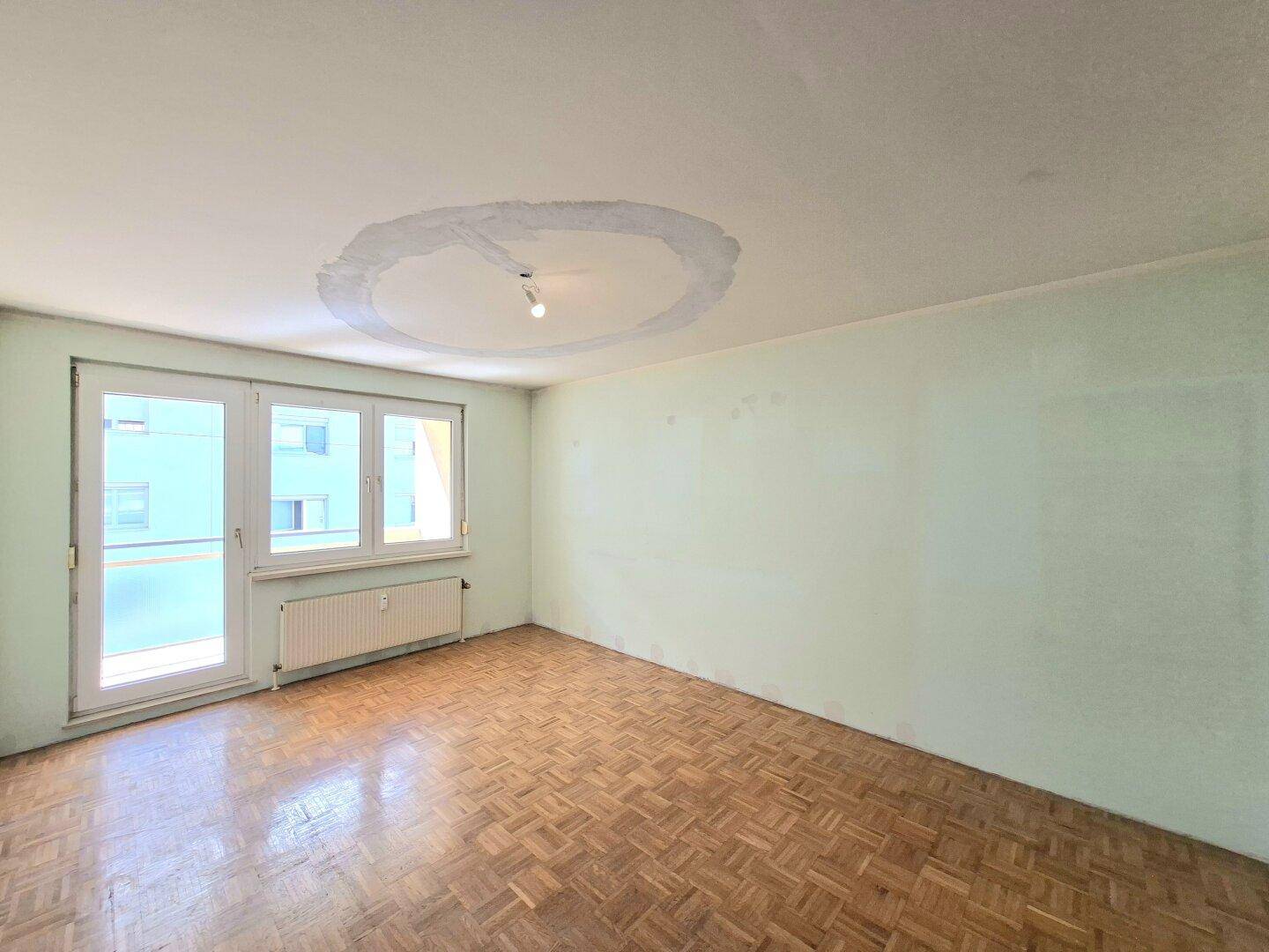 Unmöbliertes Wohnzimmer mit Fischgrätparkett, hellen Wänden und Zugang zum Balkon.