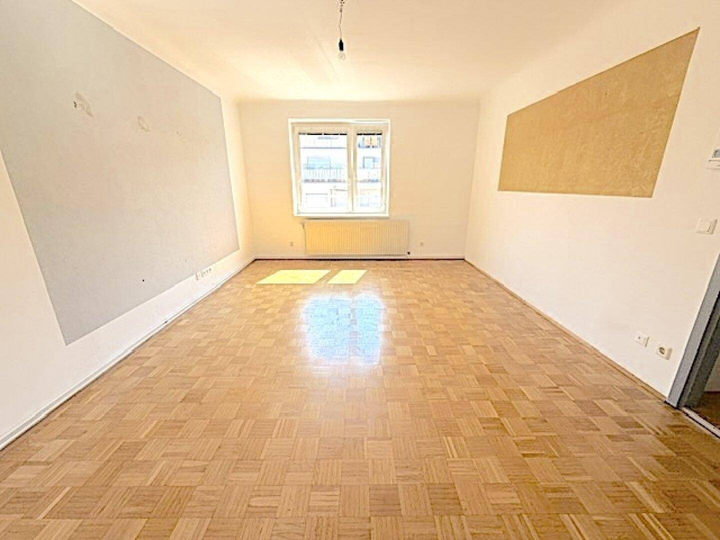 Geräumiges, leeres Wohnzimmer mit Fischgrätparkett und heller Wandgestaltung.