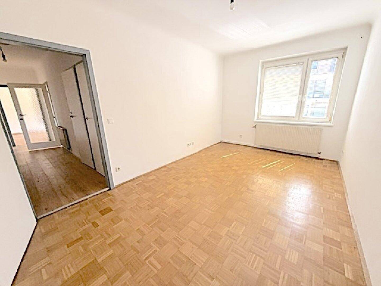 Geräumiges, leeres Wohnzimmer mit Fischgrätparkett und Zugang zum Flur.
