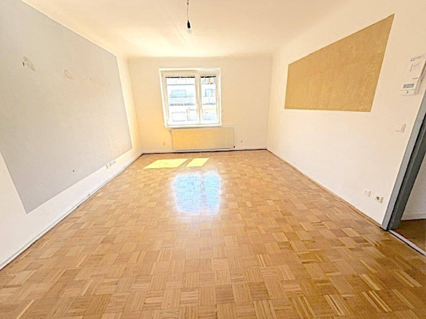 Geräumiges, leeres Wohnzimmer mit Fischgrätparkett und heller Wandgestaltung.