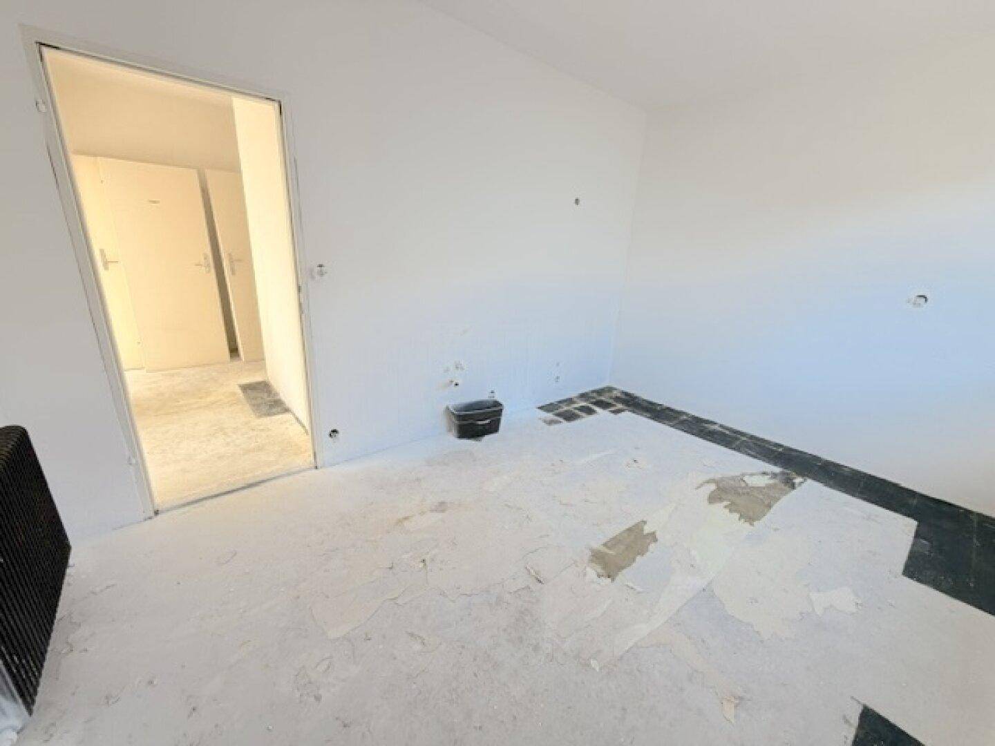 Leerer Raum mit weißen Wänden, teilweise entferntem Bodenbelag und Zugang zu einem weiteren Zimmer.
