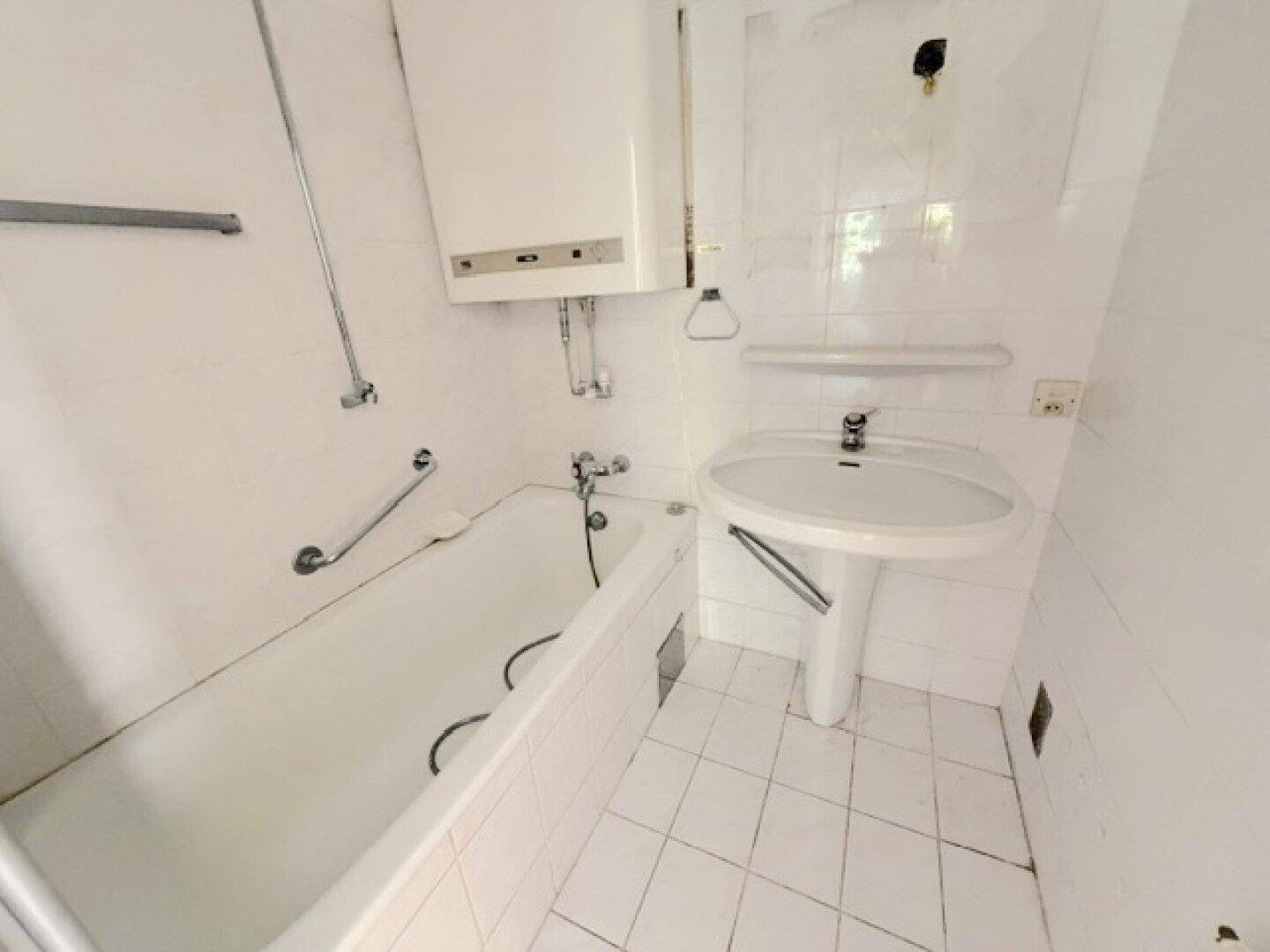 Badezimmer mit Badewanne, Waschbecken, Warmwasserbereiter und weißen Fliesen an Wänden und Boden.