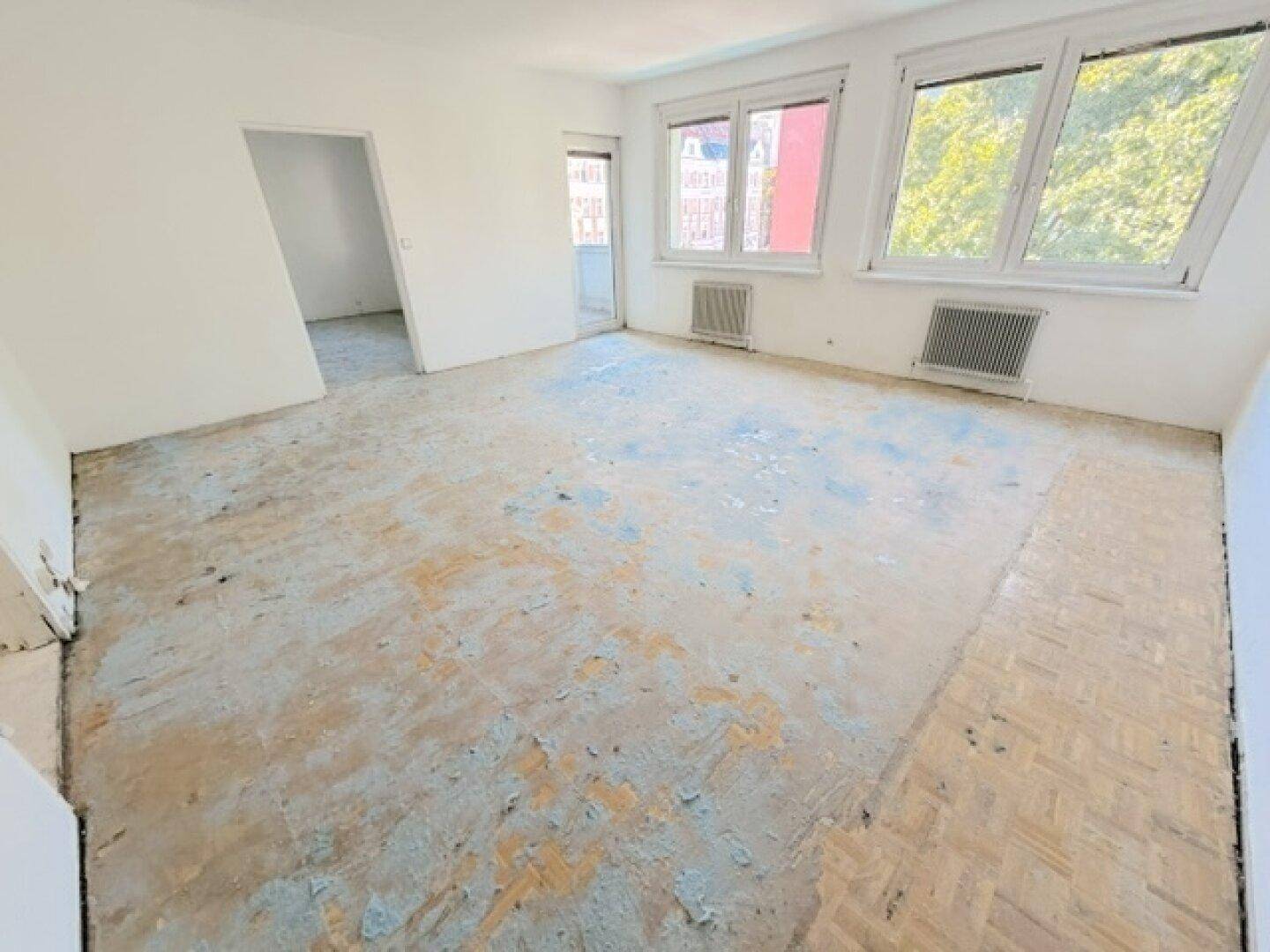 Geräumiges Zimmer mit weiß gestrichenen Wänden, altem Bodenbelag und Zugang zum Balkon.