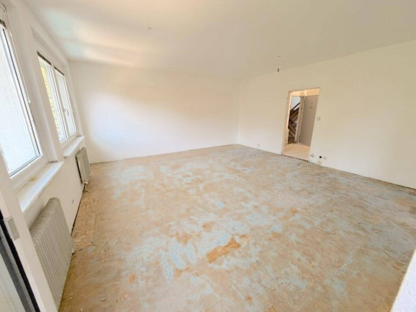 Helles Zimmer mit weiß gestrichenen Wänden, altem Bodenbelag und Zugang zu einem Treppenhaus.