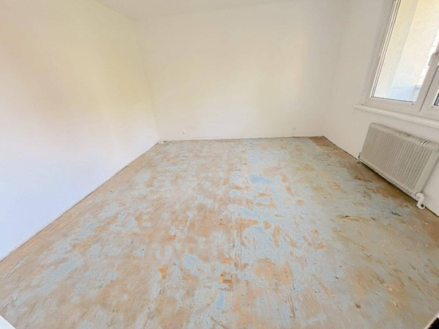 Leeres Schlafzimmer mit weiß gestrichenen Wänden, altem Bodenbelag und einem Fenster.