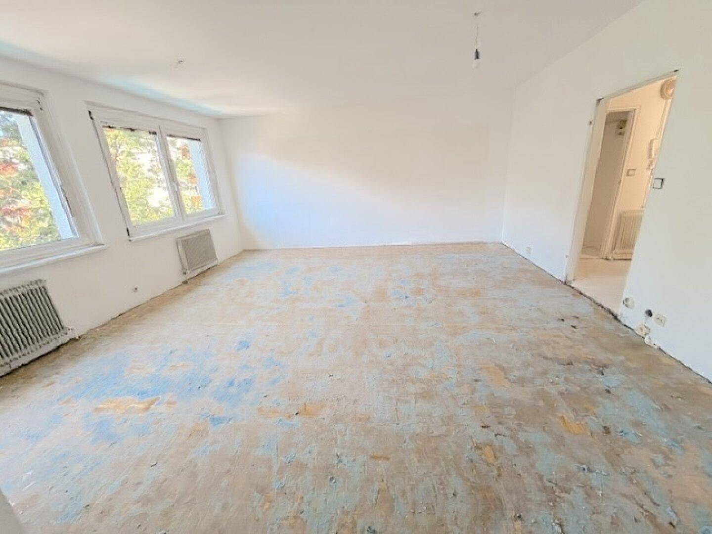 Großes Wohnzimmer mit weiß gestrichenen Wänden, altem Bodenbelag und viel Tageslicht.