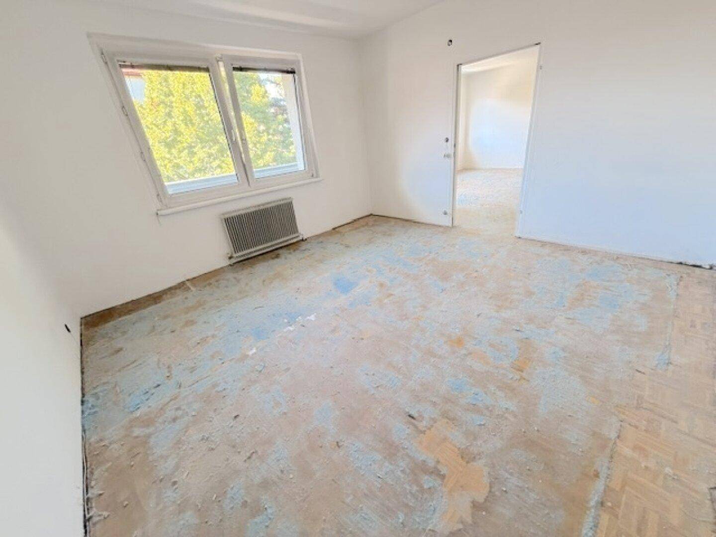 Helles Zimmer mit weiß gestrichenen Wänden, altem Bodenbelag und Blick ins Grüne.