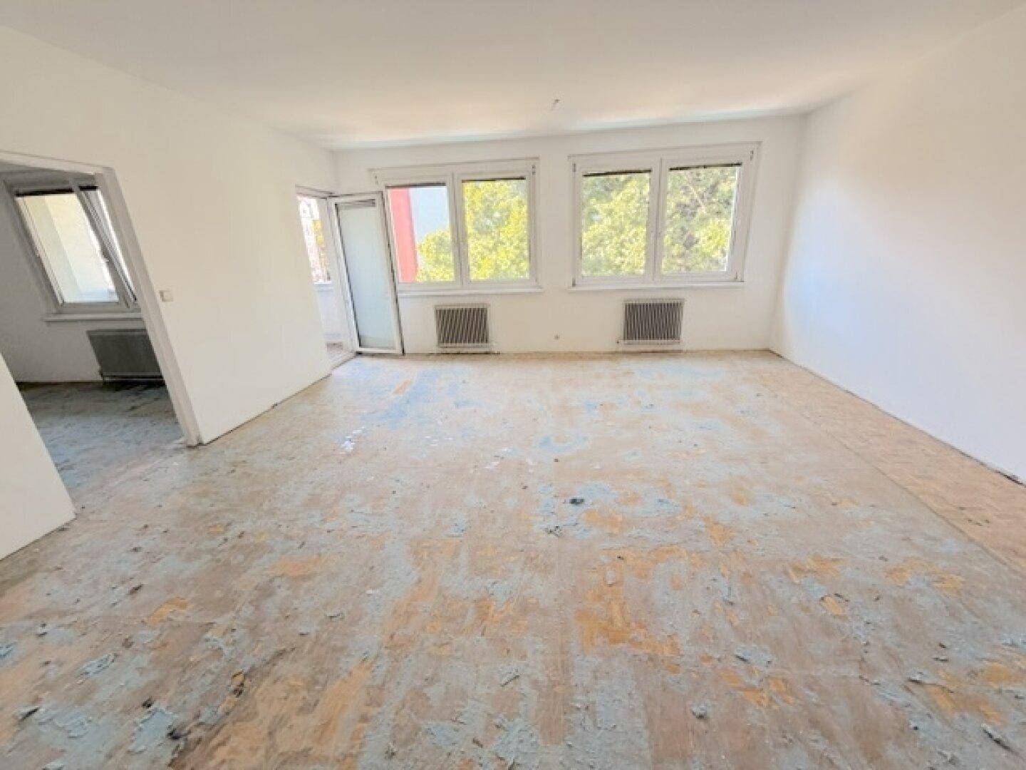 Großes Wohnzimmer mit weiß gestrichenen Wänden, altem Bodenbelag und Balkonzugang.