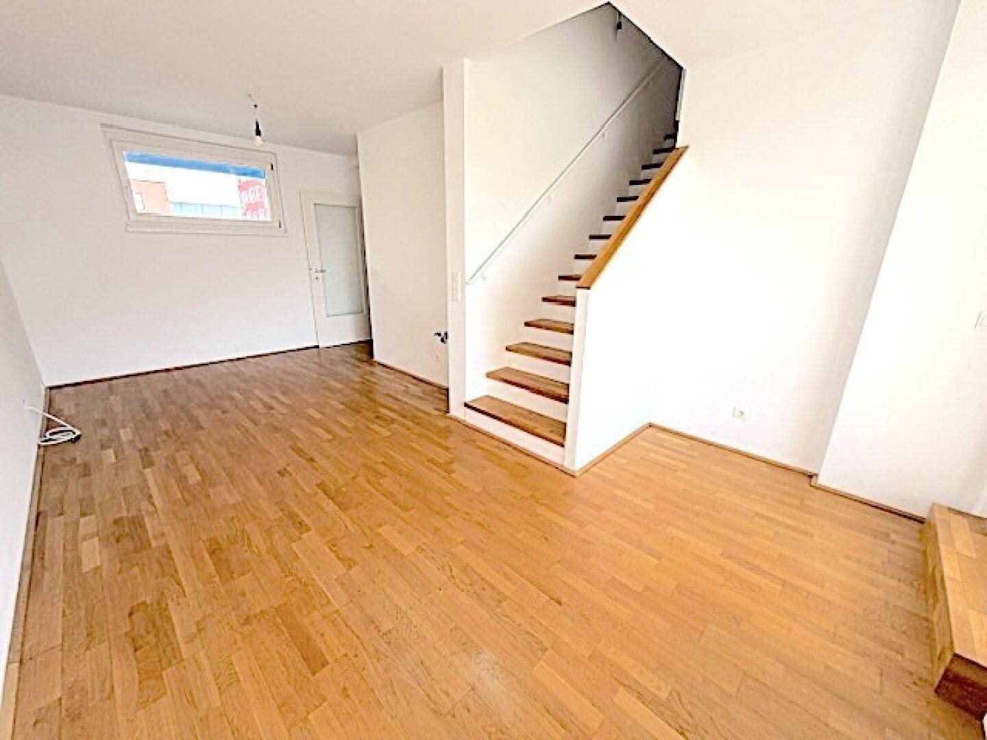 Offener Wohnbereich mit Holztreppe und großen Fenstern, die viel Tageslicht hereinlassen.