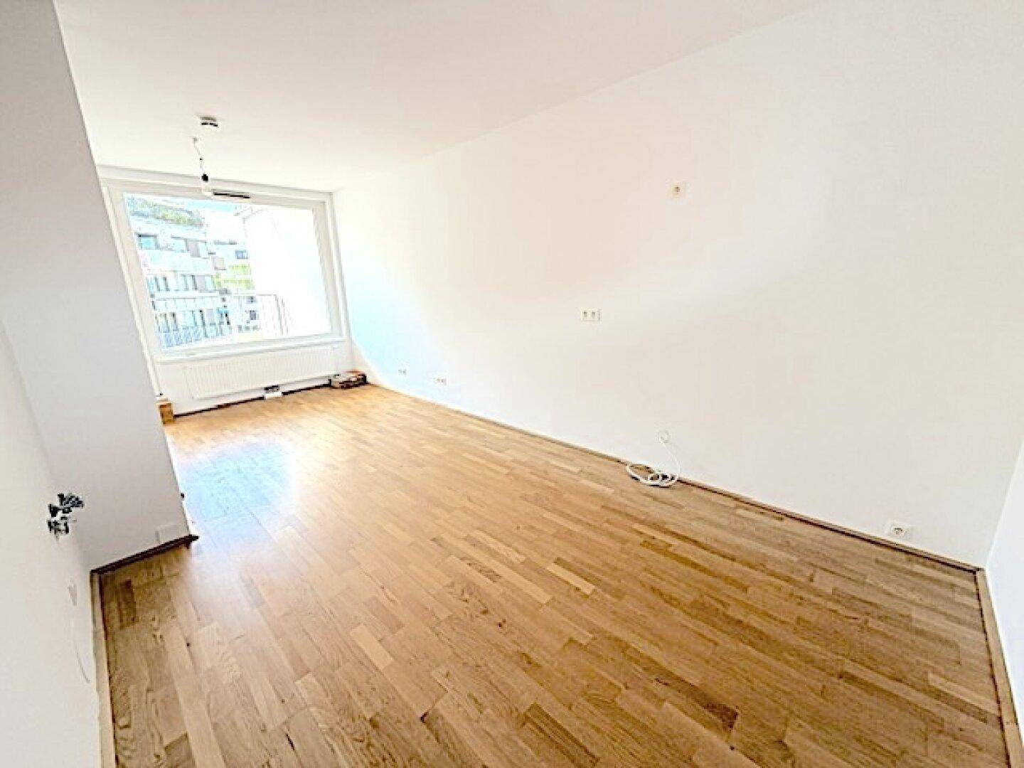 Helles und geräumiges Zimmer mit Holzboden und großem Fenster, ideal als Wohnbereich.