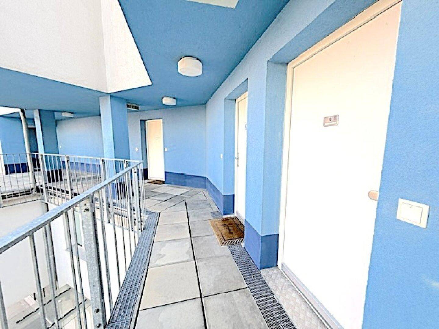 Heller Außenflur mit blauen Wänden und Fliesenboden, führt zu den Wohneinheiten.